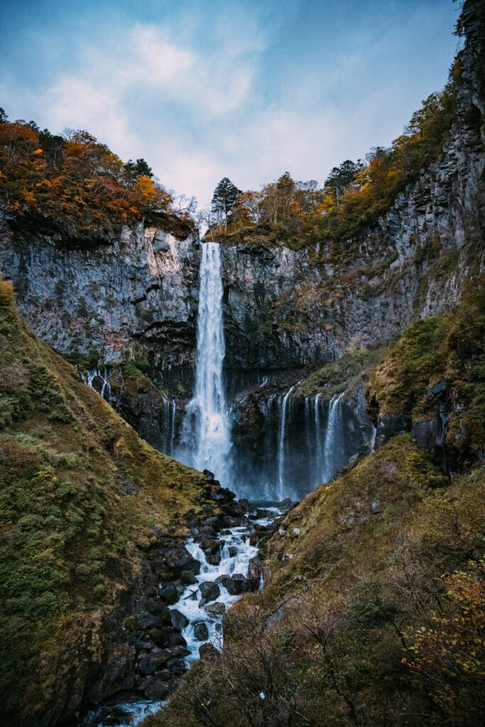 Waterfall flowing over rugged cliff with vibrant autumn leaves mossy rocks serene evening sky Wallpaper