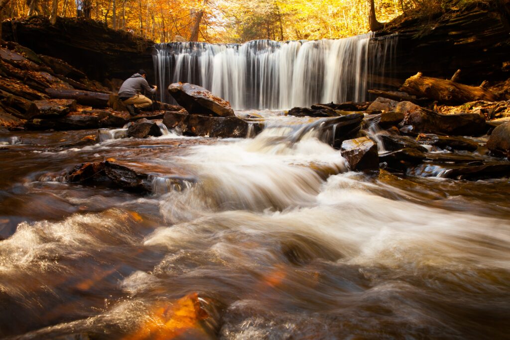 Serene autumn waterfall over dark stones with golden forest and sunlight peaceful nature scene Wallpaper