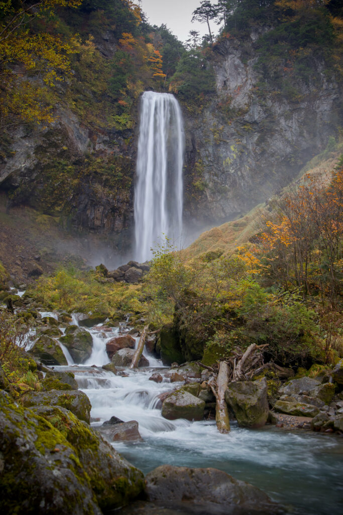Waterfall cascading over rocky cliff with autumn leaves mossy stones and clear stream flowing peacefully Wallpaper