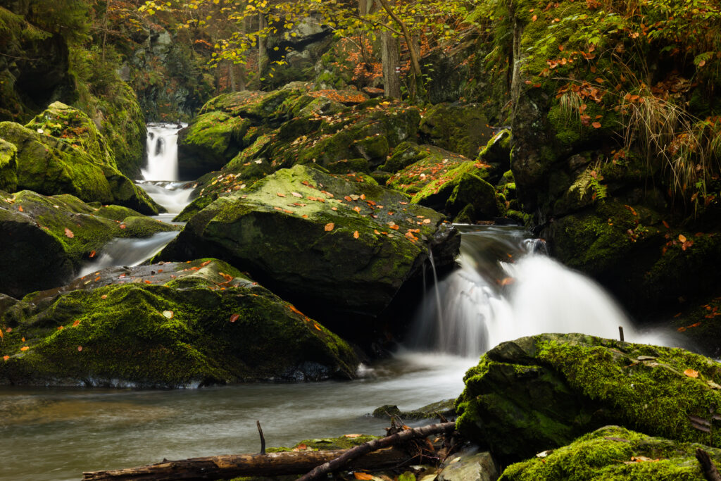 Silky waterfall flowing over mossy stones with autumn leaves in peaceful forest landscape Wallpaper