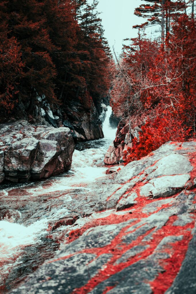 Serene waterfall flowing between rugged stones with vibrant red autumn leaves along banks Wallpaper