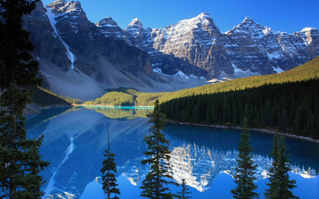 Tranquil alpine lake reflecting snow-capped mountains and pine forests in cool serene tones Wallpaper