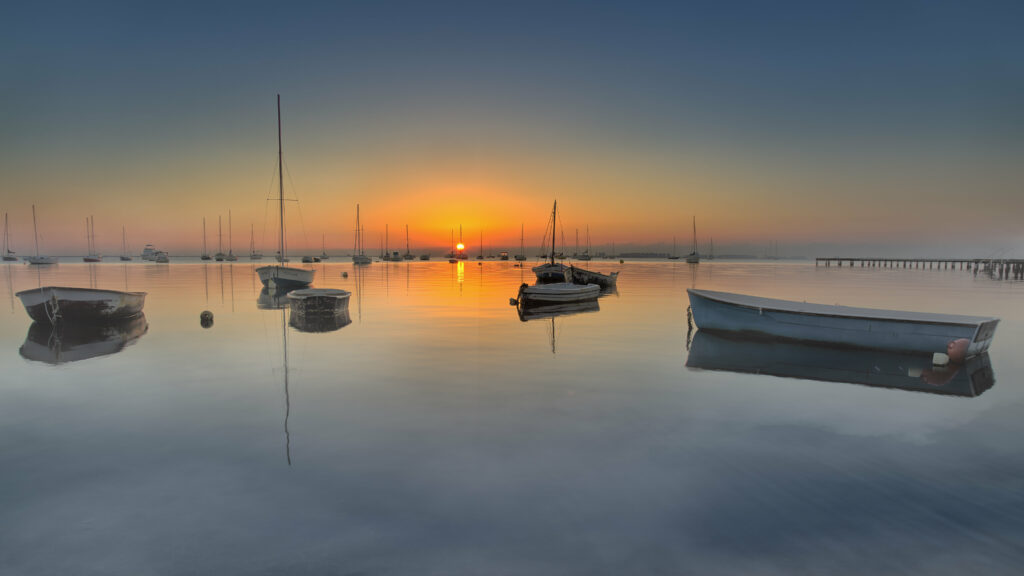 Serene sunset bay with boats floating on calm sea waters soft orange and blue reflections Wallpaper