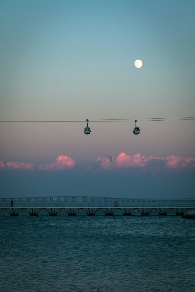 Cableway gondolas glide above a calm bridge over blue water under a pastel sky with full moon Wallpaper