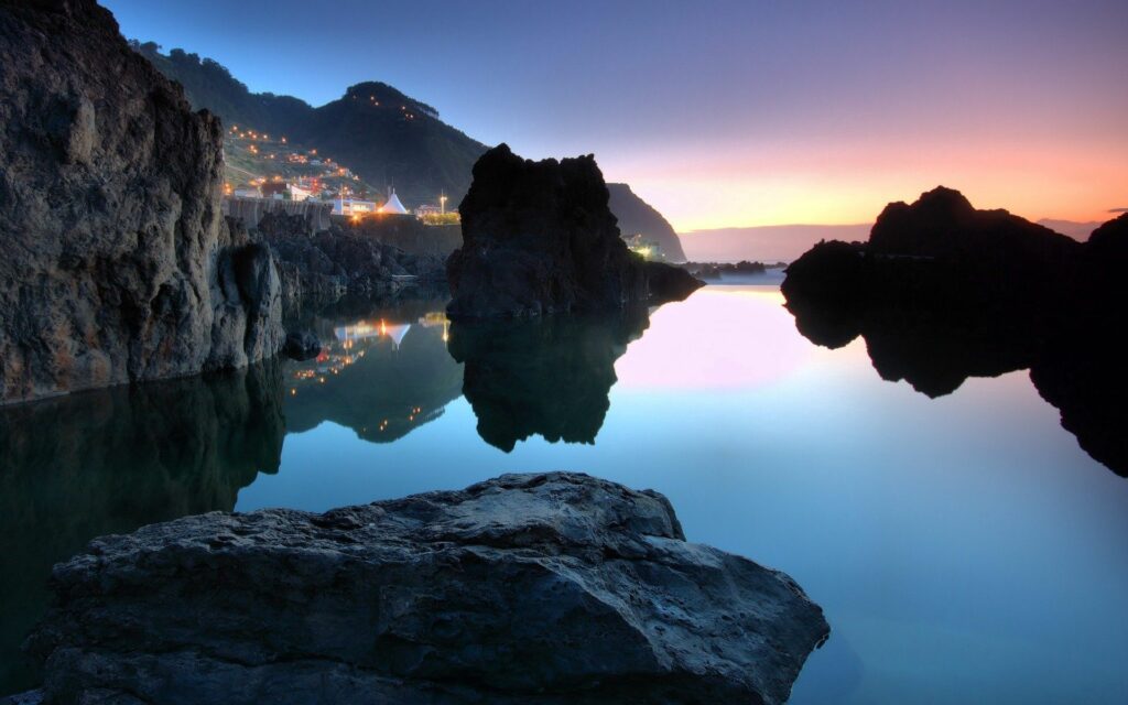 Serene evening coastal scene with rugged rocks reflected in calm water and hillside lights Wallpaper