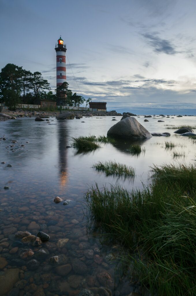 Red and white lighthouse at tranquil coastal shore with calm sea rocks grassy patches sunset sky Wallpaper