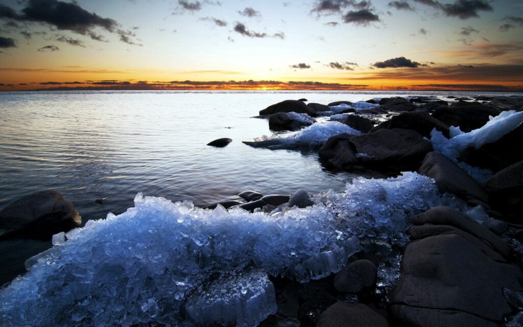 Serene coastal scene with jagged ice on rocky shores under colorful twilight sky reflecting calm sea Wallpaper