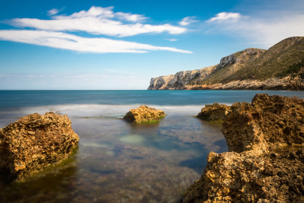 Serene coastal landscape with rugged rocks calm blue sea distant cliffs and bright cloudy sky wallpaper