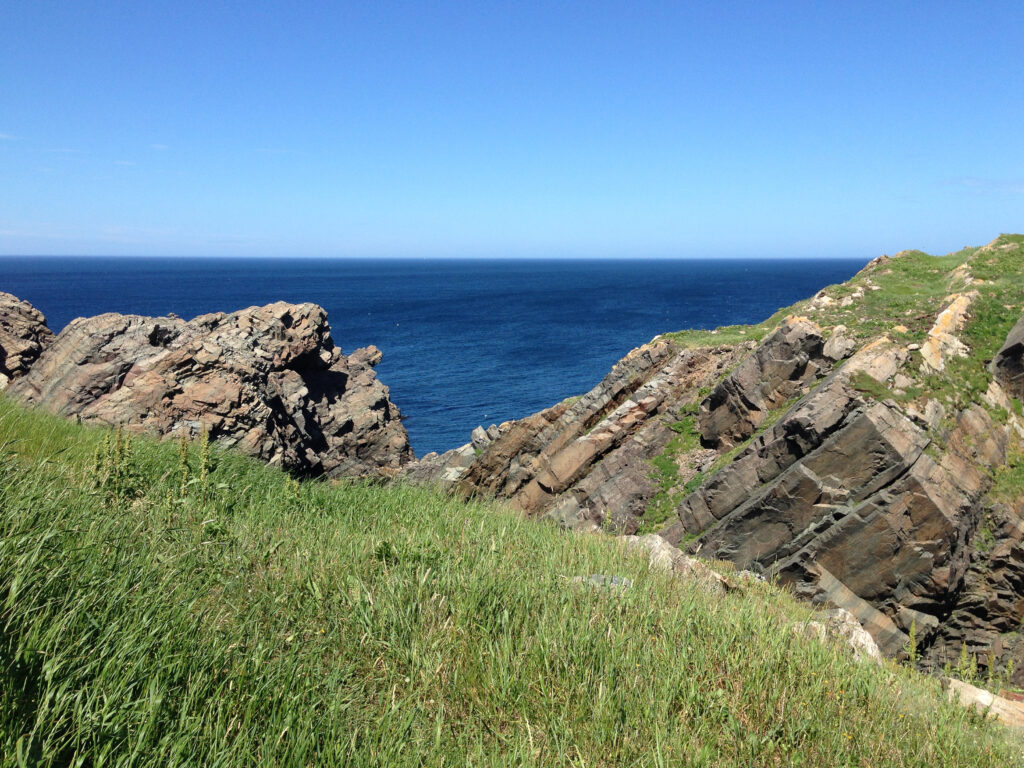 Serene coastal landscape with rugged rocks lush green grass and deep blue sea under clear sky Wallpaper