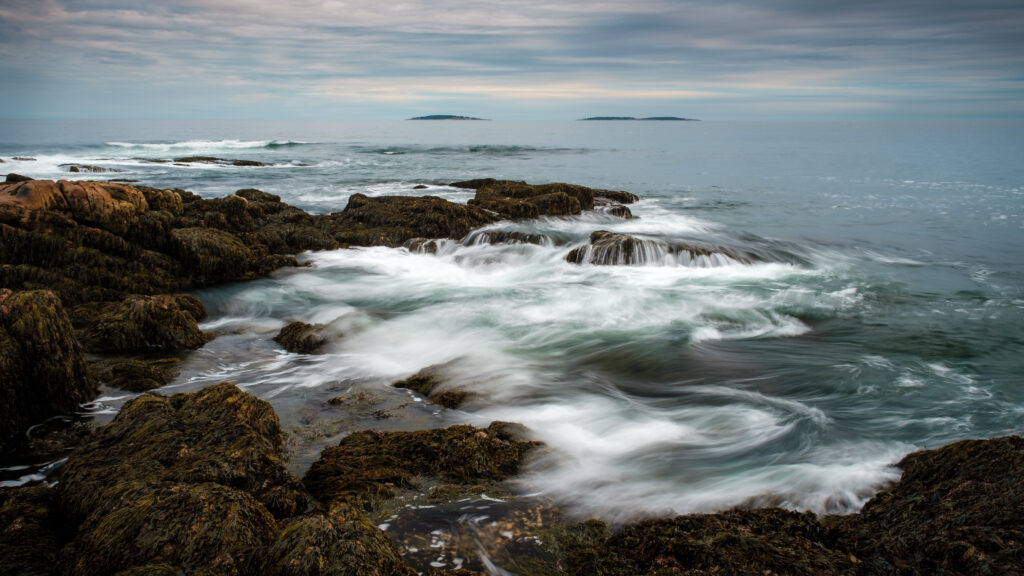 Serene coastal landscape with swirling sea waves mossy rocks distant islands and cloudy sky wallpaper