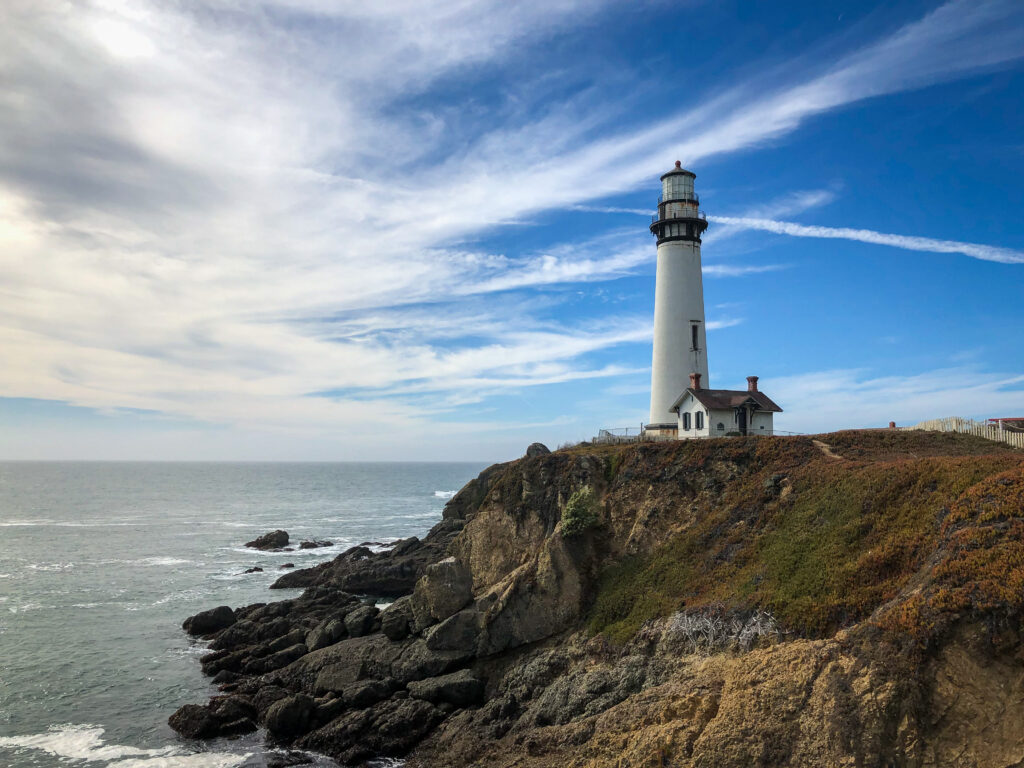 Tall white lighthouse on rocky coast overlooking vibrant blue sea and sky serene wallpaper background