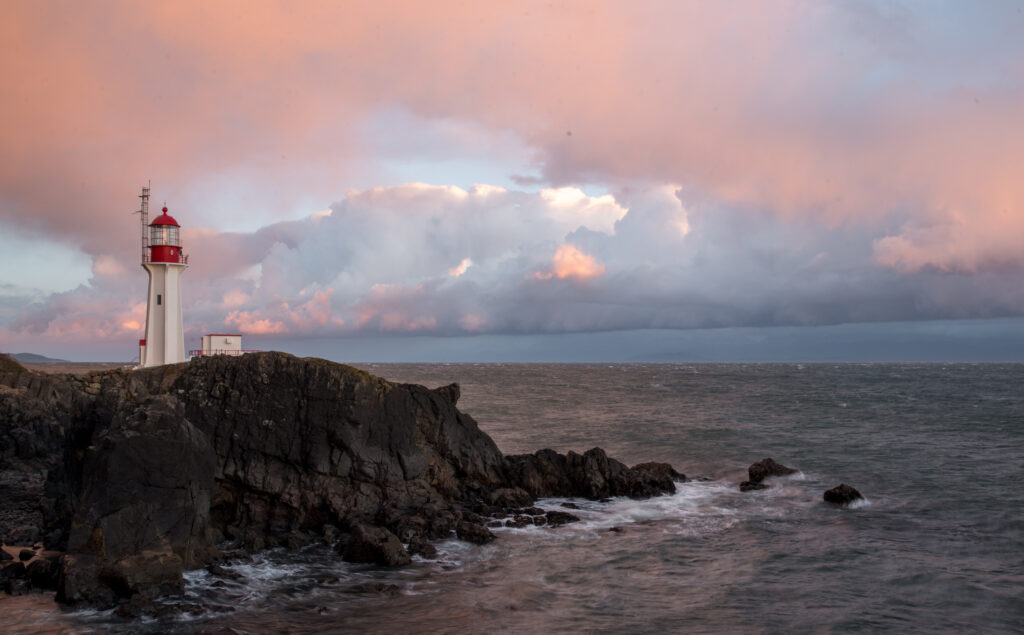 Solitary lighthouse on rocky cliffs at sunset with pink orange sky and calm coastal sea wallpaper