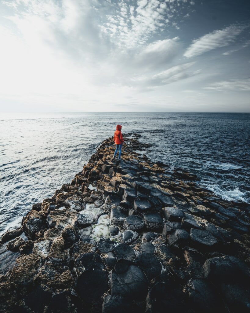 Person in red jacket on dark stone path by sea with dramatic clouds serene coastal scene wallpaper