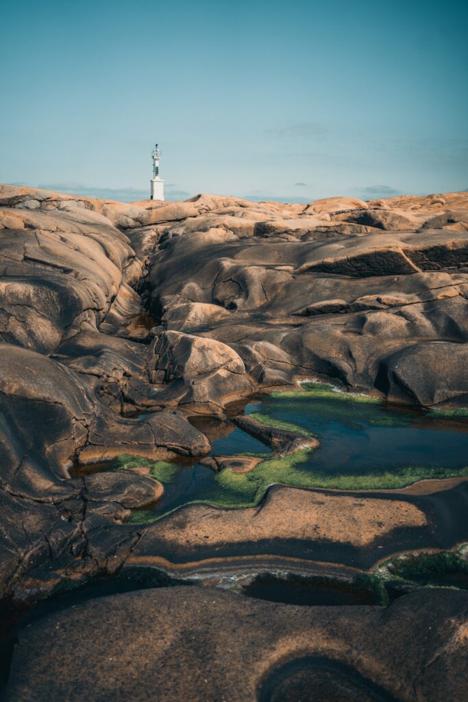 Rugged coastal rocks with shallow pools and distant lighthouse under clear blue sky wallpaper