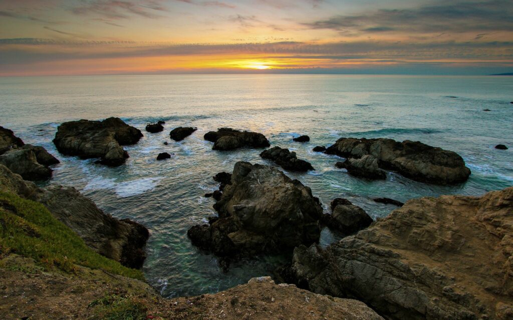 Serene coastal sunset with rugged rocks gentle waves and grassy shore peaceful wallpaper background