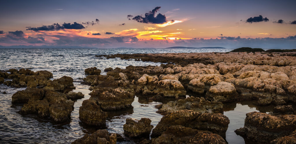Serene coastal sunset with rugged stones calm sea waves and warm orange purple sky landscape wallpaper
