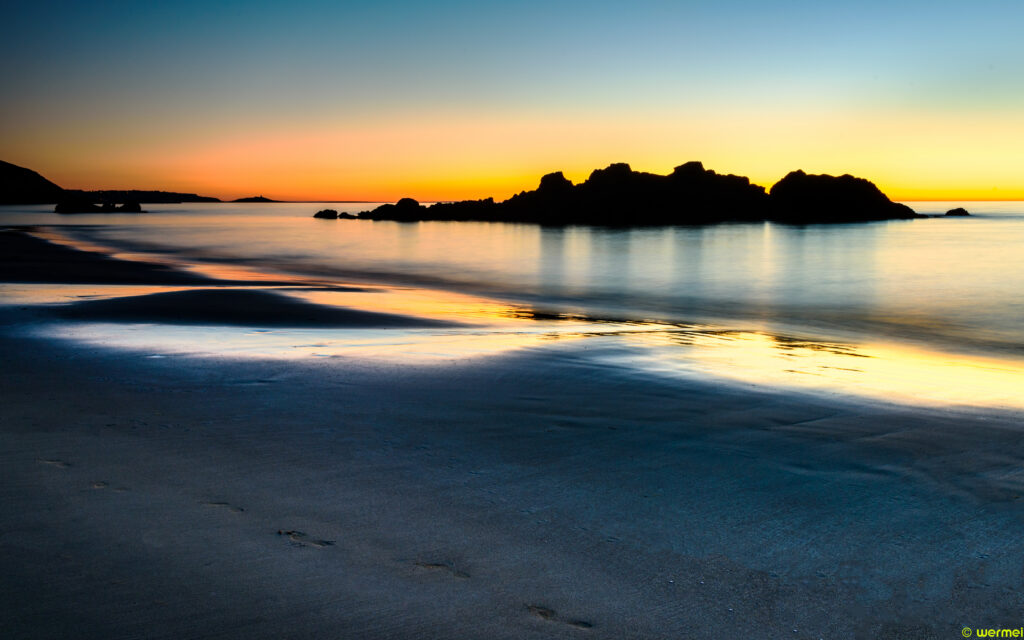 Sunset coastal scene with rocky mountains silhouetted over calm sea and sandy shore wallpaper