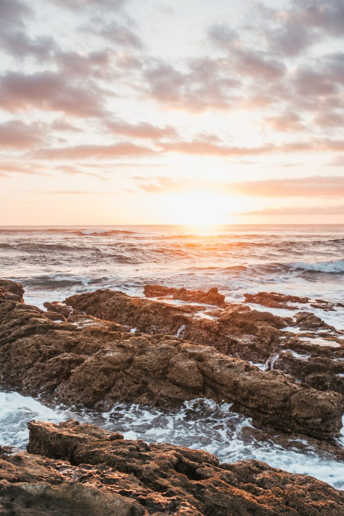 Sunset coastal wallpaper with warm sunlight on waves and rugged rocks in serene pastel sky