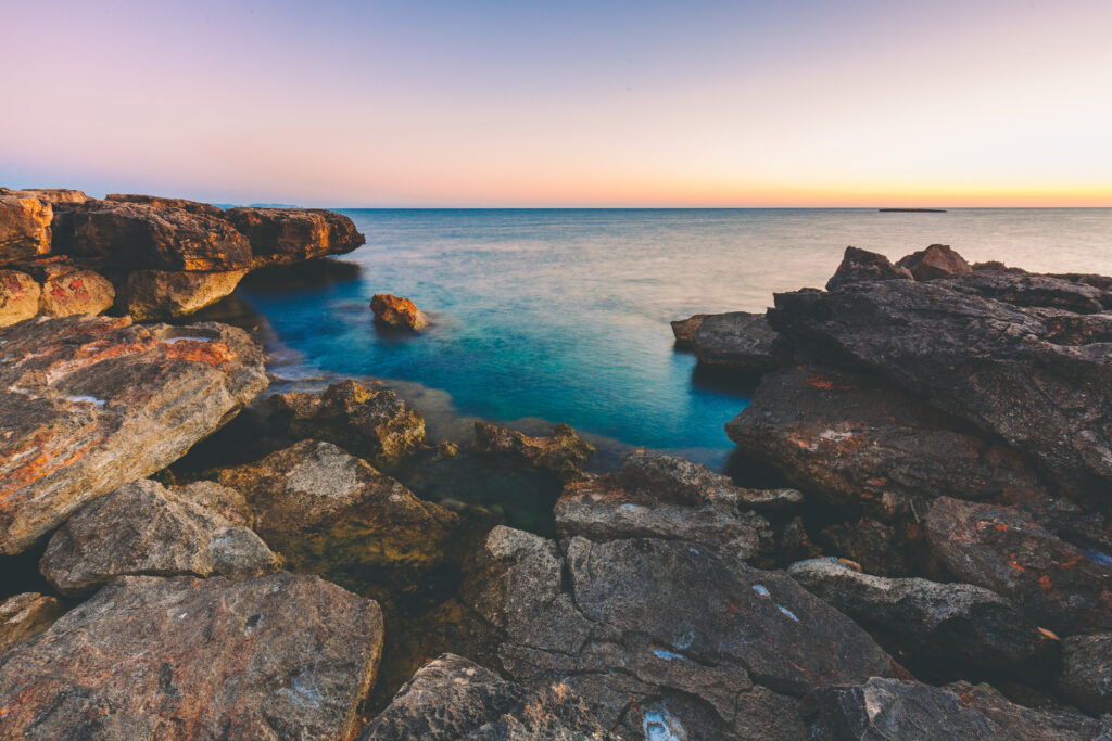 Serene coastal sunset with rugged rocks and calm blue sea in soft pastel sky wallpaper background