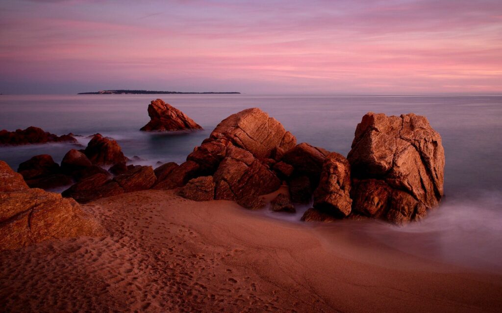 Rugged rocks on sandy beach with gentle waves and pastel sunset sky coastal wallpaper background