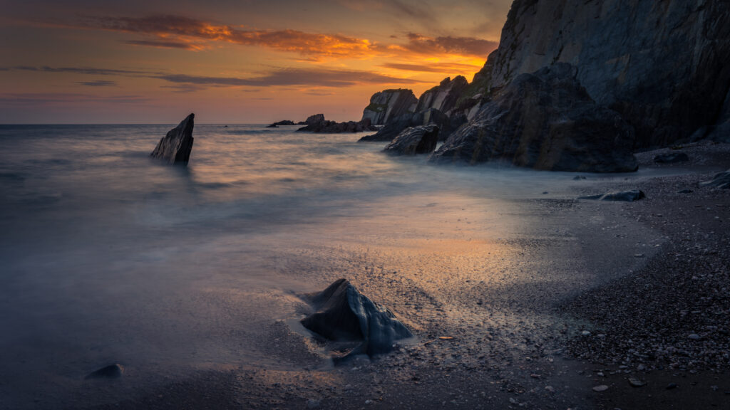 Serene sunset coastal wallpaper with rugged rocks soft sand and warm orange purple sky hues