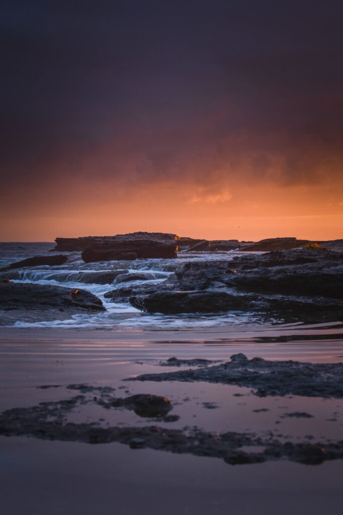 Sunset coastal shoreline with rugged rocks deep blue water and vivid orange purple sky reflection Wallpaper