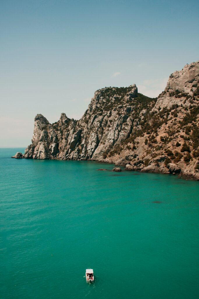 Small boat on turquoise ocean by rocky cliffs under clear blue sky serene coastal wallpaper background