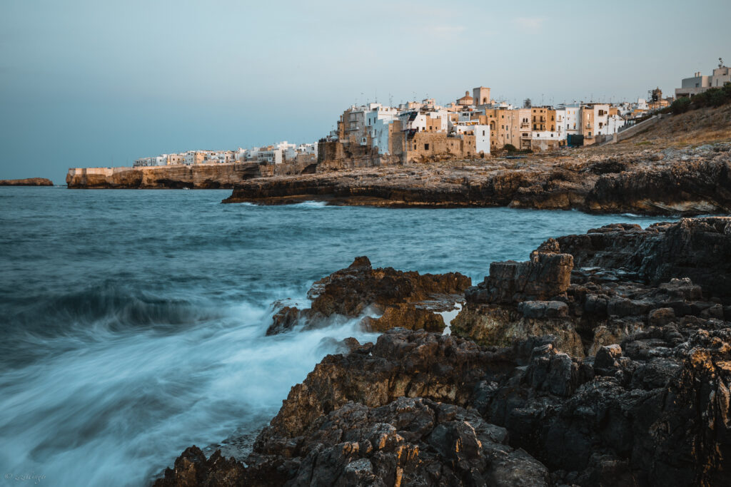 Serene coastal scene with dark rocks waves and whitewashed cliffside buildings in soft evening light Wallpaper