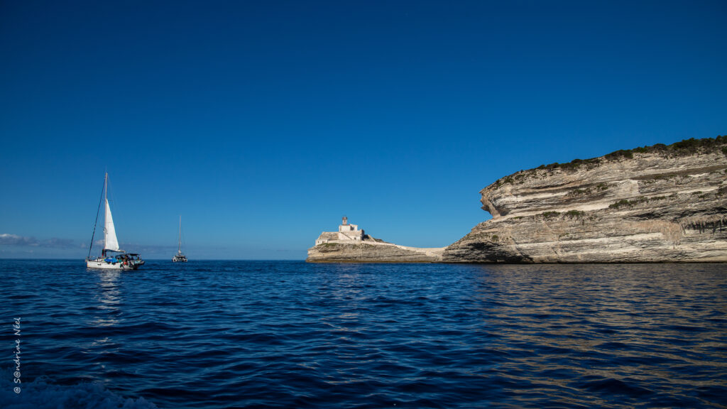Rugged rock formation and lighthouse above deep blue sea with sails in a serene coastal scene Wallpaper