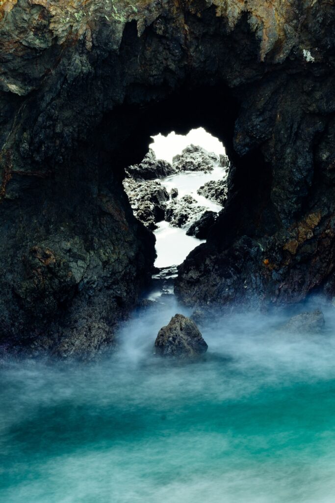 Turquoise coastal water flowing around rugged dark rocks with natural rock arch in background Wallpaper