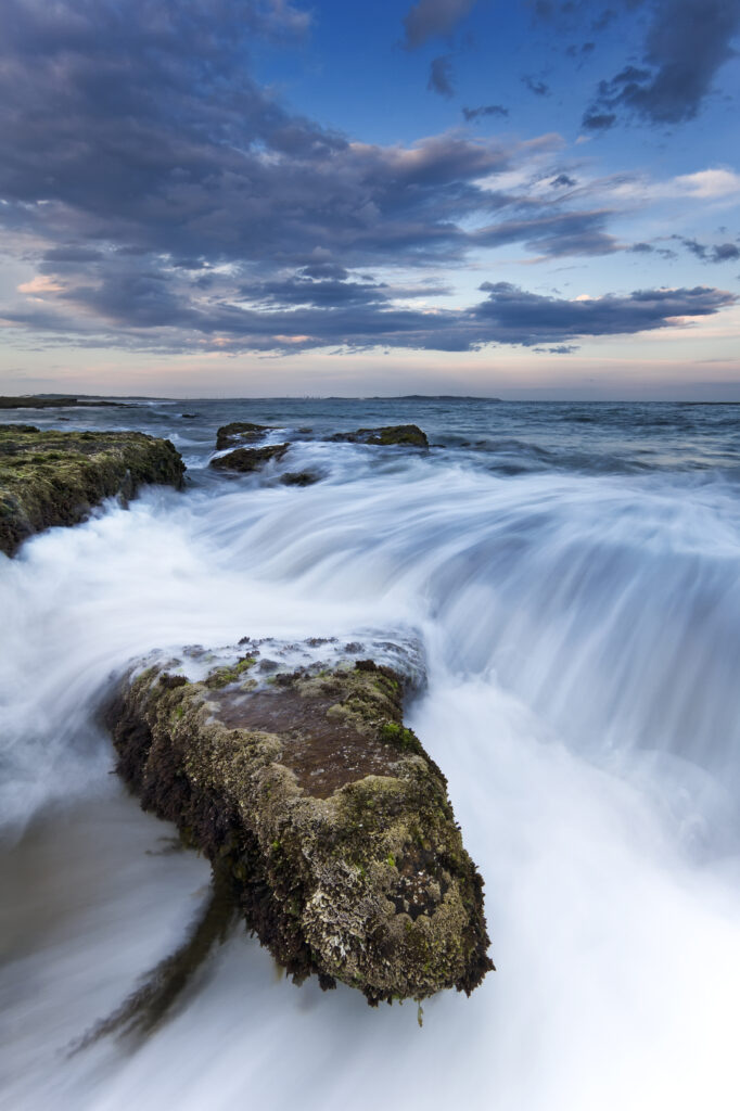 Cascading seawater over rugged stones under dramatic sky vivid blue waves coastal wallpaper background