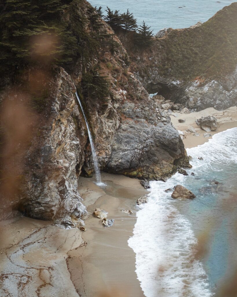 Serene coastal wallpaper with rugged rocks gentle waterfall sandy beach lush greenery calm blue sea