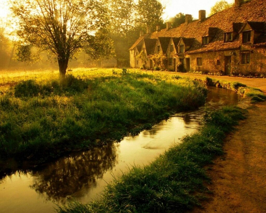 Serene morning countryside with sunlit stone buildings lush green grass and flowing tranquil channel Wallpaper