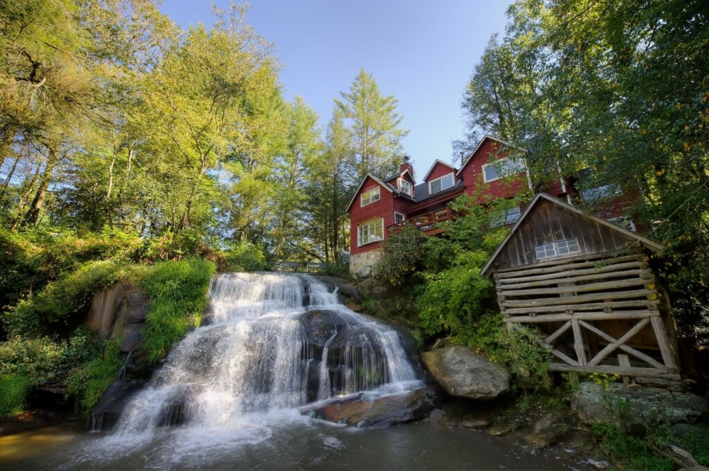 Red cottage and rustic shed by waterfall in lush green yard with sunlight through tall trees Wallpaper