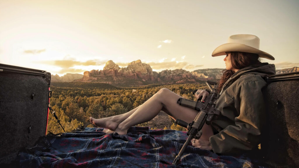Cowgirl in wide-brimmed hat resting with rifle on blanket at sunset rugged Western landscape wallpaper background