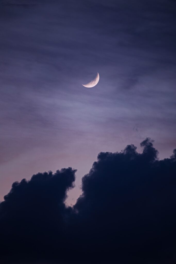 Crescent moon glowing above dark clouds in purple blue twilight sky serene wallpaper background