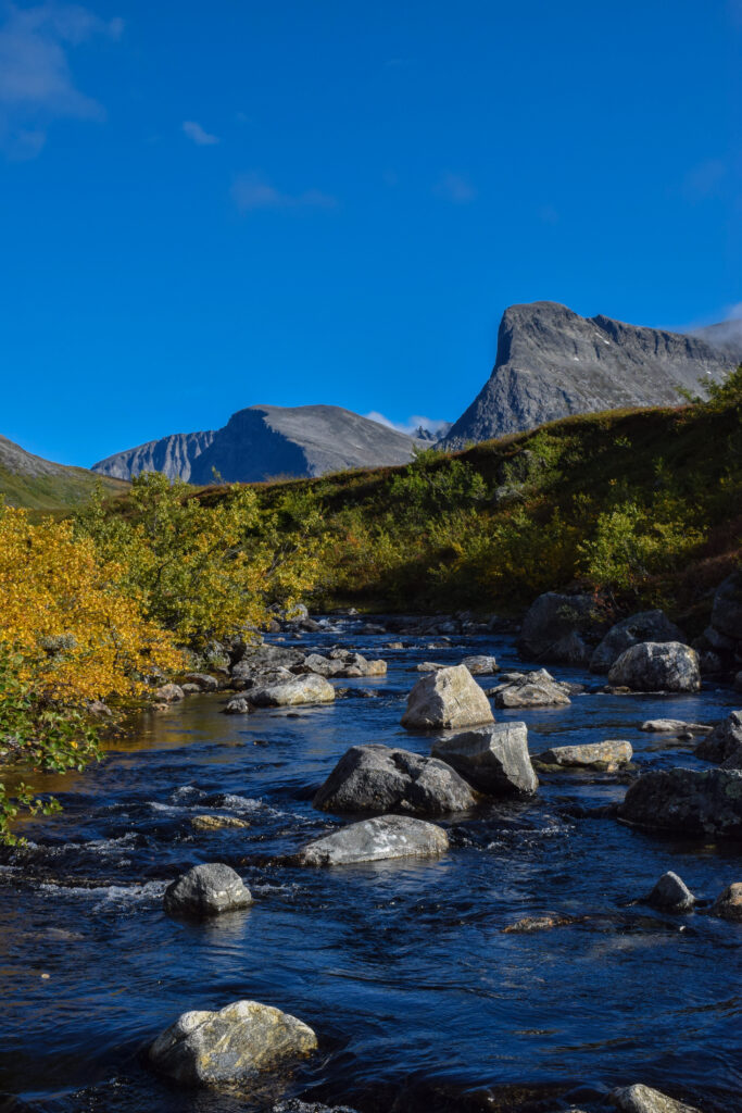 Crystal clear river over smooth stones with autumn foliage and rugged mountains under blue sky Wallpaper