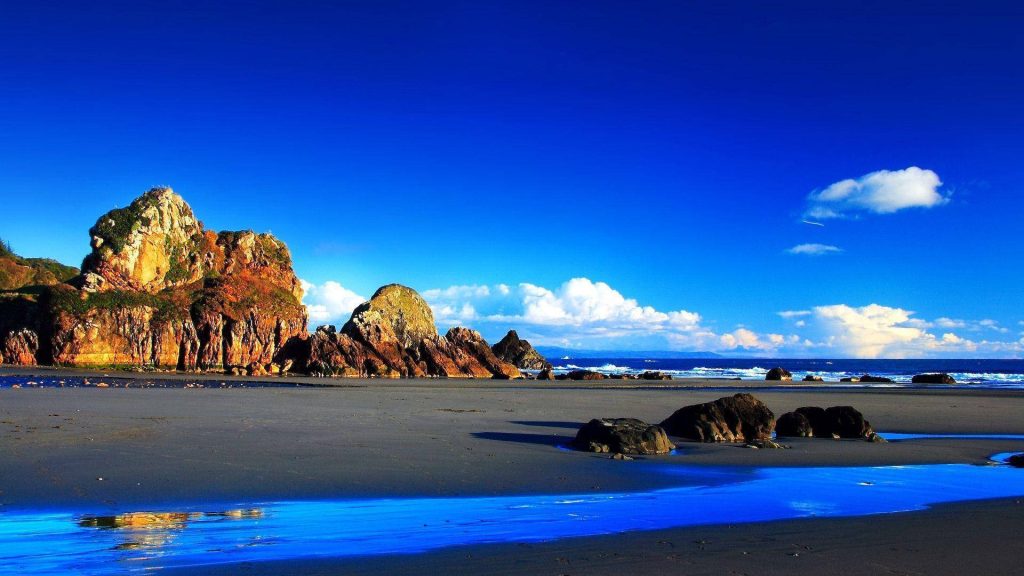 Serene dark sand beach with golden cliffs and reflective tidal pools under vibrant blue sky Wallpaper