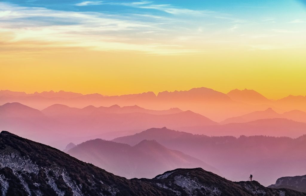 Lone hiker silhouette on rocky ridge at dawn with misty mountains and vibrant gradient sky wallpaper