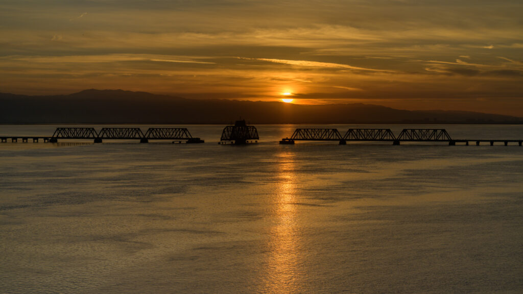 Serene dawn over sea with golden ripples and long bridge beneath dramatic cloudy sky wallpaper