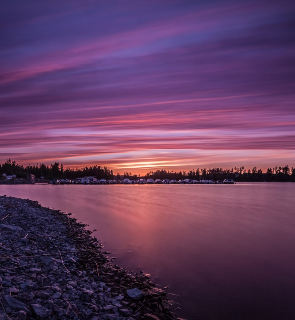 Serene river at dawn with pastel purple and pink sky reflecting on calm water and stone coast Wallpaper