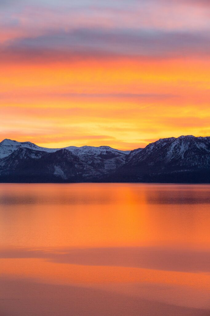 Snow-capped mountains at dawn with vibrant orange and pink sky reflected on calm sea wallpaper