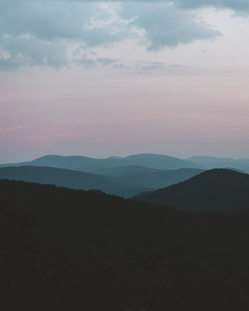 Layered mountain peaks and rolling hills in soft fog at dusk with pastel pink and blue sky Wallpaper