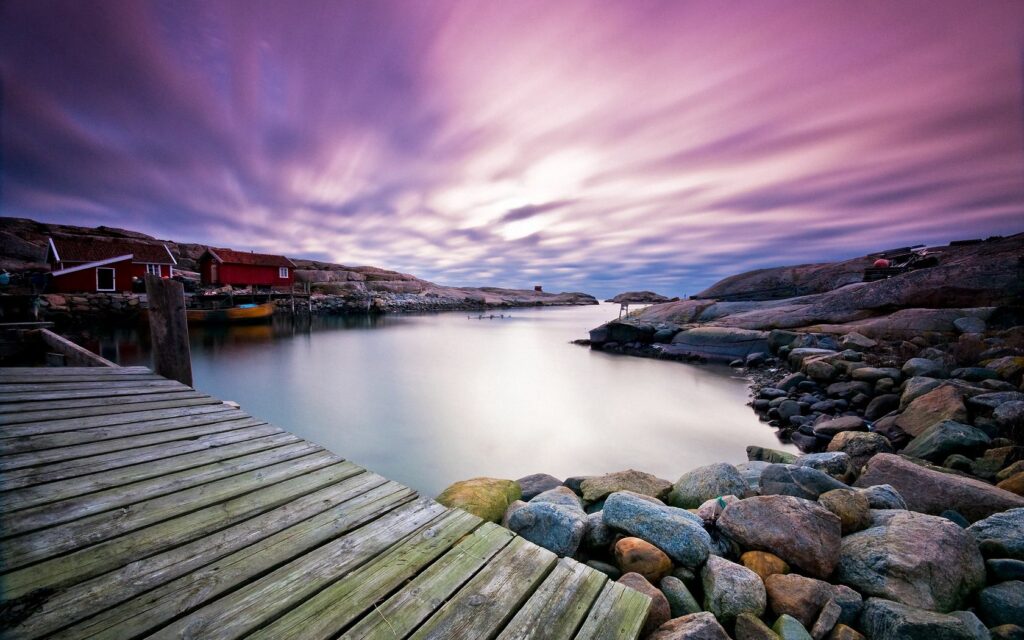 Serene evening wooden boardwalk bridge over calm water with smooth rocks and purple sunset sky Wallpaper