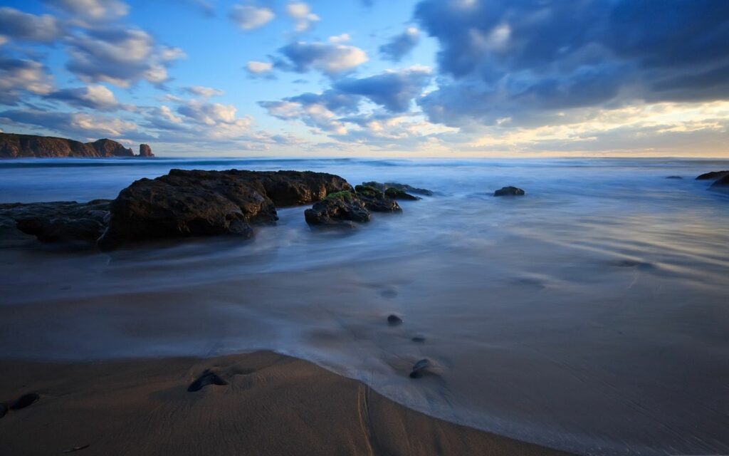 Serene evening coastal wallpaper with smooth waves sandy shores rugged stones and golden clouds