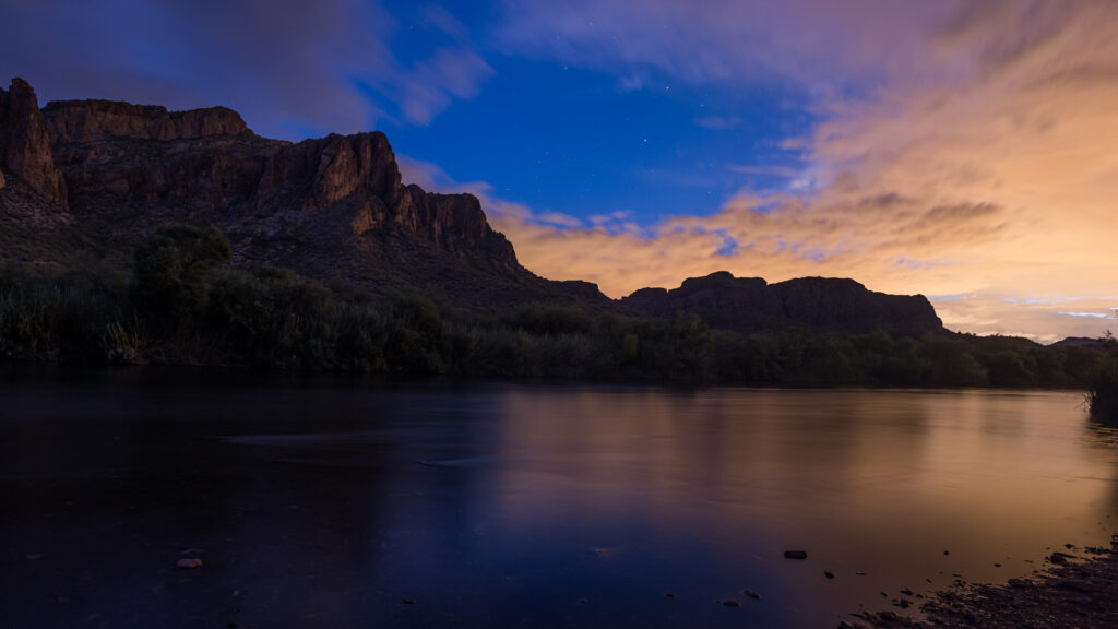 Serene evening lake with rugged rocks lush greenery and vibrant sky in landscape photography wallpaper