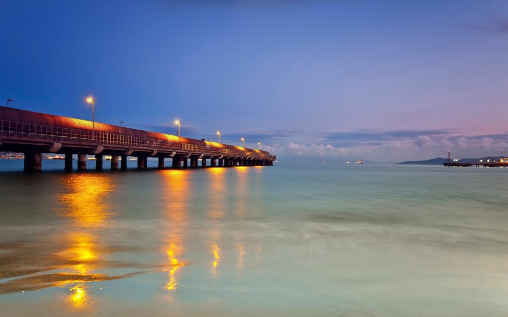 Serene evening pier over calm ocean with yellow lights reflecting under blue purple sky wallpaper