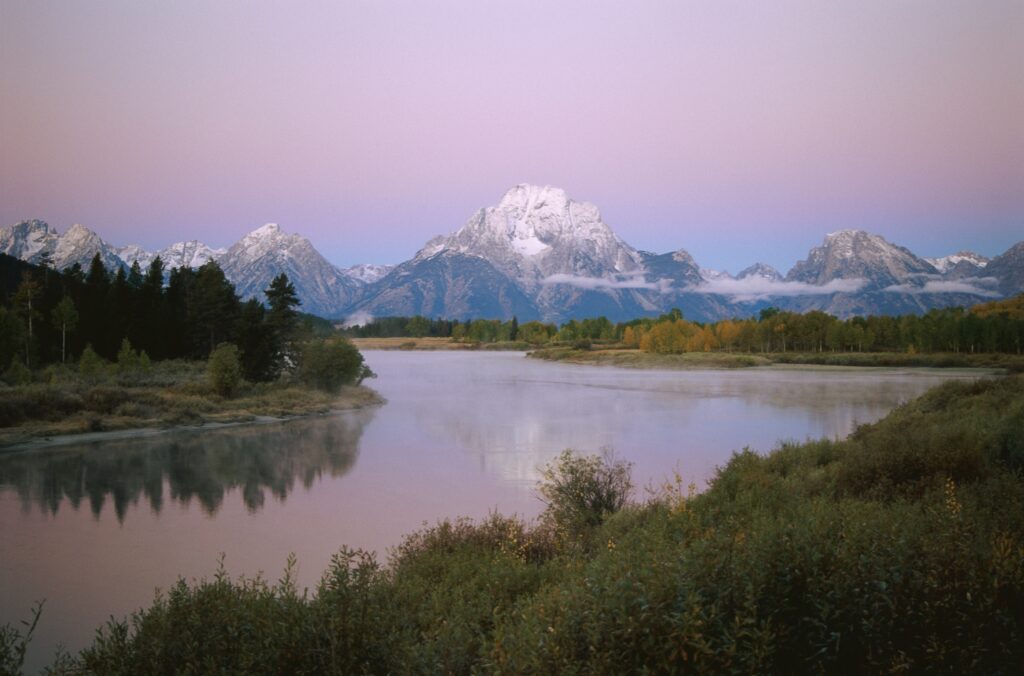 Serene evening landscape with snow-capped mountains reflecting in calm river and lush greenery Wallpaper