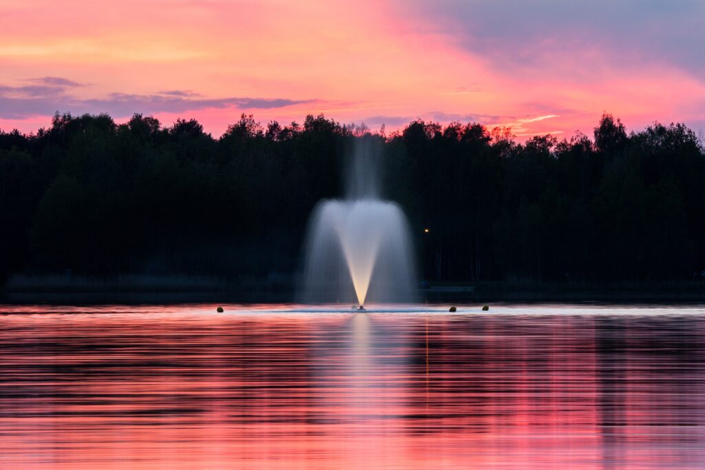 Serene fountain on calm lake at sunset with pink purple sky and silhouetted trees wallpaper background