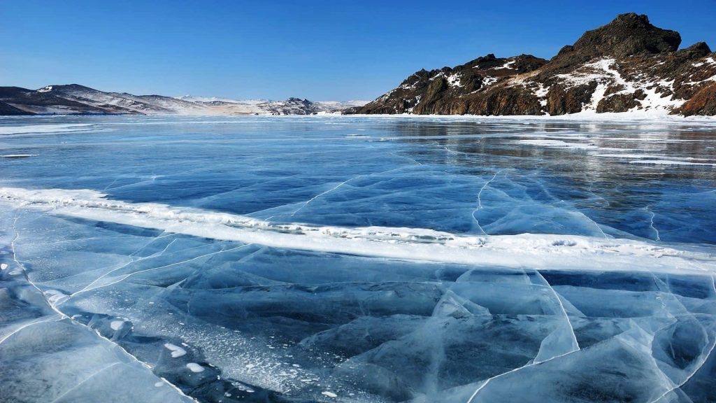 Frozen Lake Baikal with clear ice and snow-capped mountains under bright blue winter sky wallpaper
